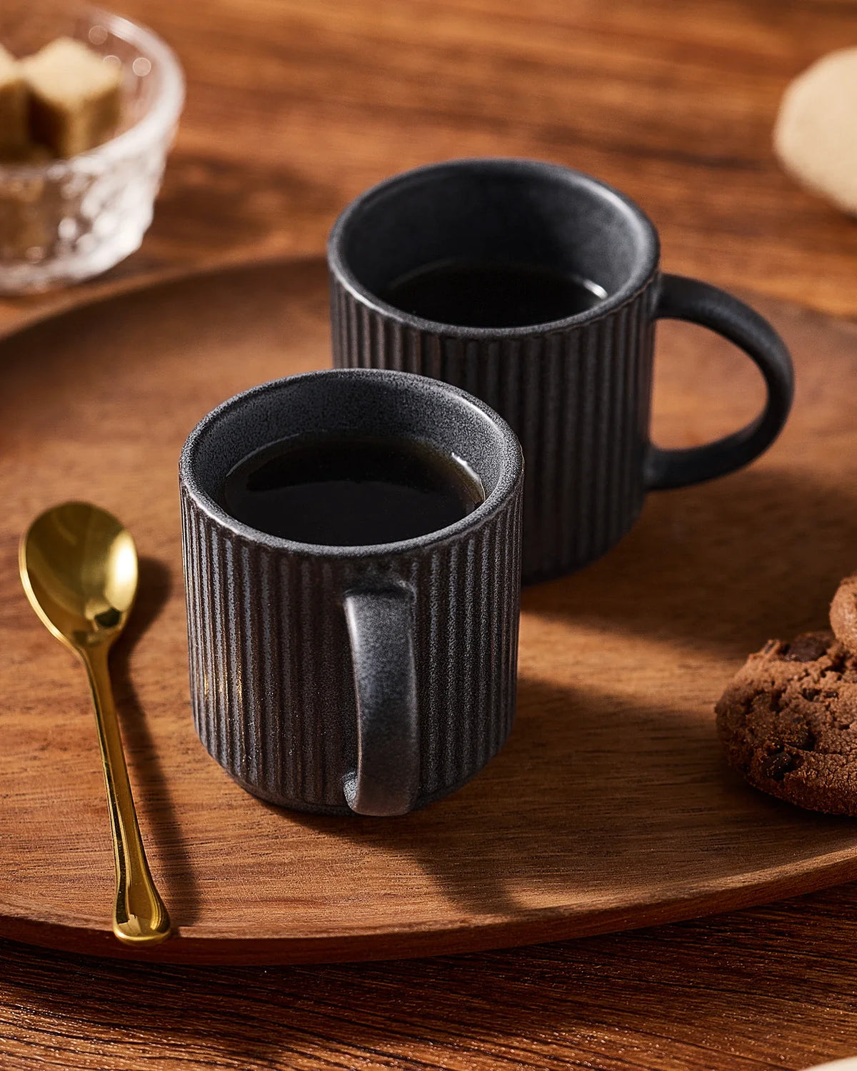 Two dark ribbed ceramic espresso cups on a wooden breakfast table.